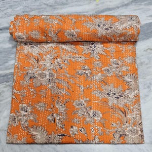 Puede incluir: Un edredón floral naranja con una sección enrollada. El edredón presenta un patrón repetido de flores y follaje beige y gris. Las costuras son visibles, agregando textura a la tela. El edredón está doblado sobre una superficie gris claro.