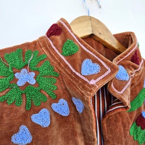 May include: A brown velvet jacket with embroidered details. The jacket features green floral and blue heart-shaped embroidery, with red and green accents on the collar. The jacket is hanging on a wooden hanger.