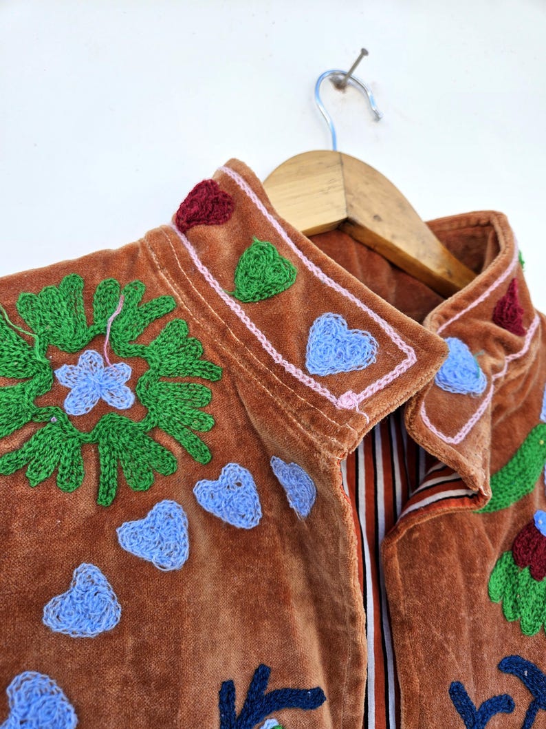 May include: A brown velvet jacket with embroidered details. The jacket features green floral and blue heart-shaped embroidery, with red and green accents on the collar. The jacket is hanging on a wooden hanger.