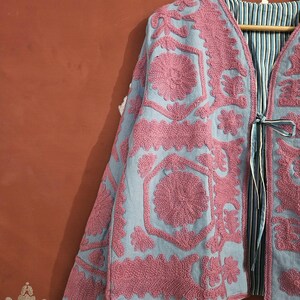 May include: A light blue jacket with intricate pink floral embroidery. The jacket features a V-neck, long sleeves, and a striped lining. It is hanging on a wooden hanger, showcasing the detailed craftsmanship and design.