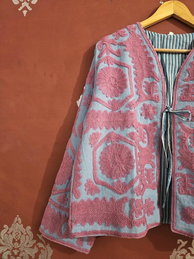 May include: A light blue jacket with intricate pink floral embroidery. The jacket features a V-neck, long sleeves, and a striped lining. It is hanging on a wooden hanger, showcasing the detailed craftsmanship and design.