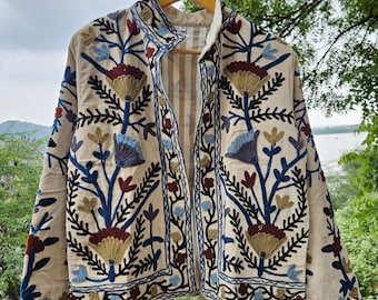 May include: Beige jacket with intricate floral embroidery in blue, red, and gold. The jacket features a stand-up collar and is displayed on a wooden hanger. The embroidery creates a striking contrast against the beige fabric, showcasing a detailed botanical design.