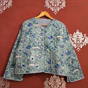 Pode incluir: Casaco acolchoado com padrão floral em tons de azul e verde. Gola redonda, dois bolsos frontais e forro listrado. Exibido em um cabide de madeira.