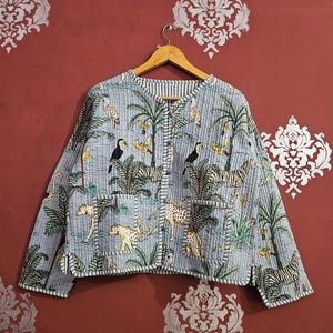 Può includere: Giacca trapuntata azzurra con stampa a tema giungla con zebre, leopardi, tucani e palme. La giacca ha un colletto rotondo, due tasche e una fodera a righe. La giacca è appesa a una gruccia di legno.