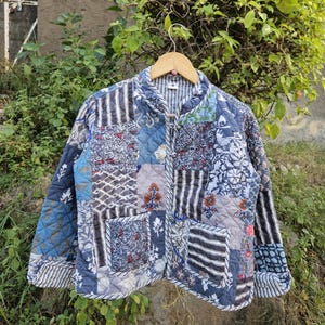 Könnte beinhalten: Gesteppte Jacke mit Patchwork-Design. Die Jacke weist verschiedene Stoffe mit Mustern in Blau, Grau und Weiß auf, darunter Blumen- und Streifenmuster. Sie hat einen runden Kragen und hängt an einem Holzbügel.