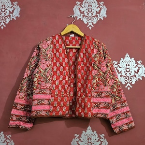 May include: A quilted jacket with a floral pattern in shades of red, pink, and brown. The jacket is open-fronted and hangs on a wooden hanger. The floral design is repeated throughout the jacket's fabric.