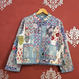 Könnte beinhalten: Gesteppte Jacke mit Patchwork-Design. Die Jacke zeigt eine Mischung aus Blumenmustern in Blau-, Rosa- und Weißtönen. Sie hat einen Stehkragen und hängt an einem Holzbügel vor rotem Hintergrund.