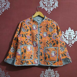 Può includere: Giacca trapuntata arancione con motivo floreale rosa, nero e bianco. La giacca ha un collo alla coreana, due tasche frontali e una chiusura con bottoni. La giacca è appesa a una gruccia di legno.