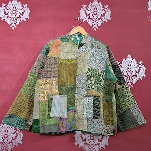 May include: A patchwork jacket in shades of green, olive, and beige, with a mandarin collar and long sleeves. The jacket features a variety of fabric patterns and textures, including floral and geometric designs. It has a single pocket on the front.
