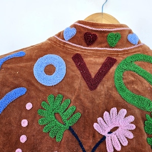 May include: Brown jacket with the word "LOVE" embroidered in blue, burgundy, and green. The jacket features embroidered flowers and hearts in shades of pink, green, and blue. The jacket is hanging on a wooden hanger.