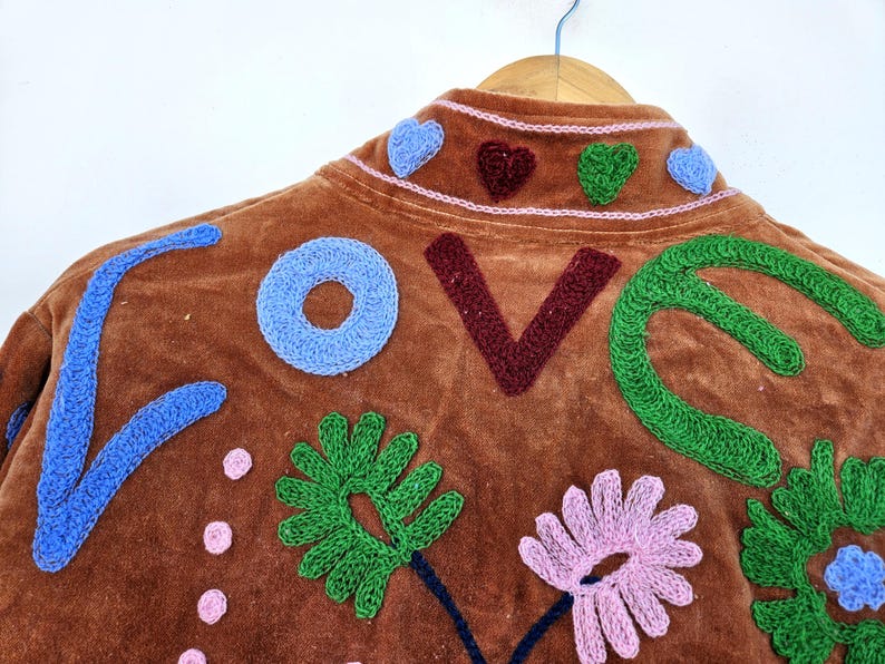 May include: Brown jacket with the word "LOVE" embroidered in blue, burgundy, and green. The jacket features embroidered flowers and hearts in shades of pink, green, and blue. The jacket is hanging on a wooden hanger.