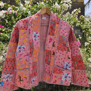 May include: A quilted jacket featuring a patchwork design with various floral and tiger motifs in shades of pink, orange, and red. The garment has long sleeves, two front pockets, a shawl collar, and a striped inner lining with a size M tag.