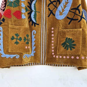 以下が含まれることがあります： 赤、青、緑の花と葉のデザインが刺繍されたマスタード色のジャケット。 ジャケットには、2つのフロントポケットと垂直ジッパーが付いています。 刺繍には、ハート、花、その他の装飾要素が含まれています。