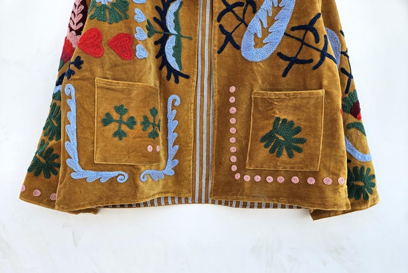 以下が含まれることがあります： 赤、青、緑の花と葉のデザインが刺繍されたマスタード色のジャケット。 ジャケットには、2つのフロントポケットと垂直ジッパーが付いています。 刺繍には、ハート、花、その他の装飾要素が含まれています。