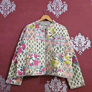 May include: A patchwork quilted jacket with a stand-up collar. The jacket displays floral and geometric patterns in pink, green, and blue on a cream base. It has two front pockets and is suspended from a wooden hanger. The jacket's design is a mix of patterns.