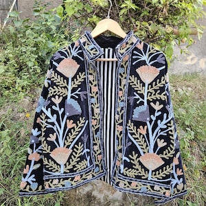 Peut inclure: Veste en velours noir avec broderies florales aux tons pêche, bleu et or. La veste a une doublure rayée noire et blanche et est présentée sur un cintre en bois. La broderie représente des fleurs et des feuilles stylisées.