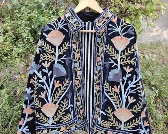 May include: A black velvet jacket with floral embroidery in shades of peach, blue, and gold. The jacket has a striped black and white lining and is displayed on a wooden hanger. The embroidery features stylized flowers and leaves.