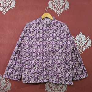 May include: A quilted jacket with a repeating geometric pattern in shades of purple, white, and cream. The jacket is hanging on a wooden hanger against a maroon background. The jacket has long sleeves and a mandarin collar.
