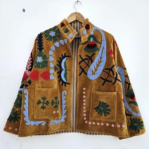 Könnte beinhalten: Senffarbene Samtjacke mit bunten gestickten Blumen- und Paisley-Designs. Die Jacke hat zwei Vordertaschen und ein gestreiftes Futter. Die Stickerei umfasst rote, blaue, grüne und rosa Akzente.