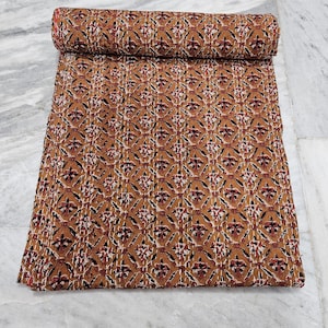 May include: A rolled-up, patterned quilt with a repeating floral design in shades of brown, red, and white. The quilt is laid out on a light-colored surface, showcasing its intricate pattern and texture. The quilt is a warm, inviting textile.
