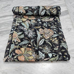 Puede incluir: Colcha floral negra con una sección enrollada. La colcha presenta un patrón detallado de flores y hojas en tonos melocotón, verde y blanco sobre un fondo negro. Las costuras son visibles, agregando textura al diseño.
