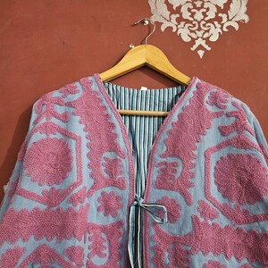 May include: A light blue jacket with intricate pink embroidery. The jacket features a V-neck and a tie closure. The interior lining is striped in blue and white. The jacket is hanging on a wooden hanger.