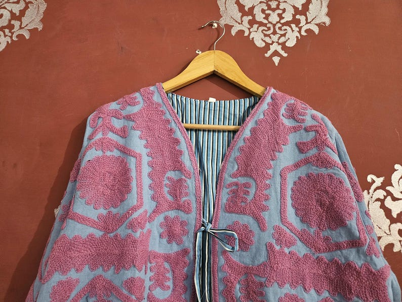 May include: A light blue jacket with intricate pink embroidery. The jacket features a V-neck and a tie closure. The interior lining is striped in blue and white. The jacket is hanging on a wooden hanger.