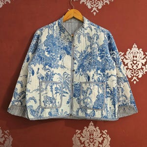 Może przedstawiać: Pikowana kurtka z niebiesko-białym wzorem toile de Jouy przedstawiającym ptaki i liście. Kurtka ma kołnierz mandaryński, dwie kieszenie i paski. Zawieszona na drewnianym wieszaku na czerwonym tle.