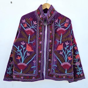 Pode incluir: Casaco de veludo roxo com bordados florais intrincados em tons de rosa, coral e azul. O casaco apresenta uma gola alta e um padrão de riscas verticais no centro. Pendurado num cabide de madeira.