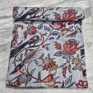 Puede incluir: Un edredón enrollado de color azul claro con un detallado estampado de pájaros y flores. El diseño presenta pájaros, flores rojas y hojas verdes sobre un fondo azul claro. El edredón está hecho de un material suave y es adecuado para una cama o un sofá.