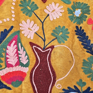 以下が含まれることがあります： マスタード色の織物で、刺繍された花柄と抽象的なデザインが施されています。濃い赤色の花瓶にはピンクと緑の花が入っています。「LOVE」の文字は青と濃い赤色の糸で刺繍されています。その他の刺繍要素には、葉や花柄が含まれています。