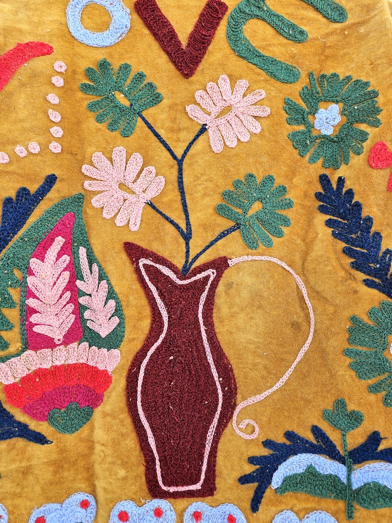 以下が含まれることがあります： マスタード色の織物で、刺繍された花柄と抽象的なデザインが施されています。濃い赤色の花瓶にはピンクと緑の花が入っています。「LOVE」の文字は青と濃い赤色の糸で刺繍されています。その他の刺繍要素には、葉や花柄が含まれています。
