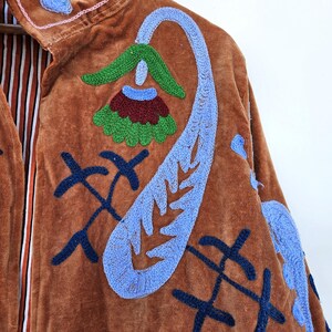 May include: A brown velvet jacket with colourful embroidered floral and abstract designs. The jacket has a high collar and a striped lining. The embroidery includes blue, green, red, and pink accents, creating a bohemian aesthetic.