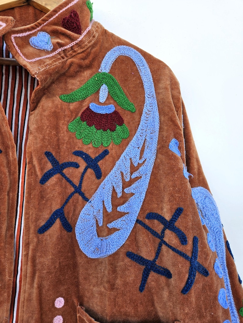 May include: A brown velvet jacket with colourful embroidered floral and abstract designs. The jacket has a high collar and a striped lining. The embroidery includes blue, green, red, and pink accents, creating a bohemian aesthetic.