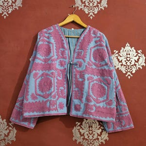 May include: A light blue jacket with pink floral embroidery. The jacket has long sleeves and a tie closure. The lining is striped. The jacket is hanging on a wooden hanger against a red wall with decorative patterns.