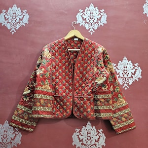 Könnte beinhalten: Eine gesteppte rote Jacke mit Blumenmuster und zwei Vordertaschen. Die Jacke hängt an einem Holzbügel vor einer roten Wand mit weißen dekorativen Akzenten. Die Jacke hat ein Rautenmuster.