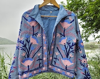 May include: A light blue jacket with intricate floral embroidery in pink and blue. The jacket is hanging on a wooden hanger against a backdrop of a lake and greenery. The embroidery design features stylized flowers and leaves.