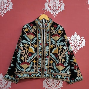Peut inclure: Veste noire brodée de motifs floraux colorés. La veste a un col montant et une doublure rayée. La broderie comprend des fleurs et des feuilles rouges, jaunes et bleues. La veste est suspendue à un cintre en bois.