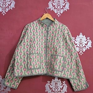 Peut inclure: Veste matelassée à motifs floraux. Le fond est vert clair avec des fleurs roses et des feuilles vertes. Elle présente une bordure rayée noire et blanche et deux poches avant. La veste est suspendue à un cintre en bois.