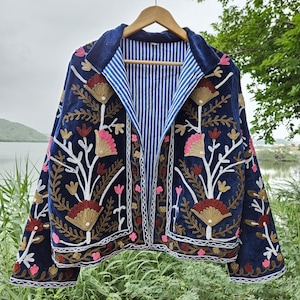 Può includere: Giacca ricamata blu navy con un design floreale. La giacca presenta una fodera a righe blu e bianca e un colletto in velluto. Il ricamo include motivi floreali rosa, oro e bianchi. La giacca è appesa a una gruccia di legno.