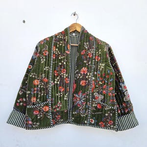 Può includere: Giacca trapuntata verde oliva con motivo floreale rosso, arancione e blu. La giacca presenta finiture a righe bianche e nere sui bordi, tasche e polsini. È appesa a una gruccia di legno.