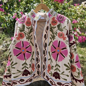 Peut inclure: Veste beige brodée avec un motif floral. La veste présente de grandes fleurs roses et oranges, des feuilles vertes et des accents bleus et marron. La veste est suspendue à un cintre en bois sur fond de fleurs.