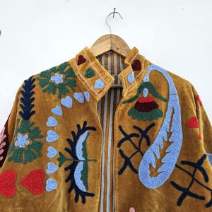 以下が含まれることがあります： マスタード色のジャケットで、複雑な刺繍が施されています。 ジャケットはハイカラーで、花、ハート、抽象的な形など、カラフルな刺繍デザインで飾られています。 裏地は縦縞です。