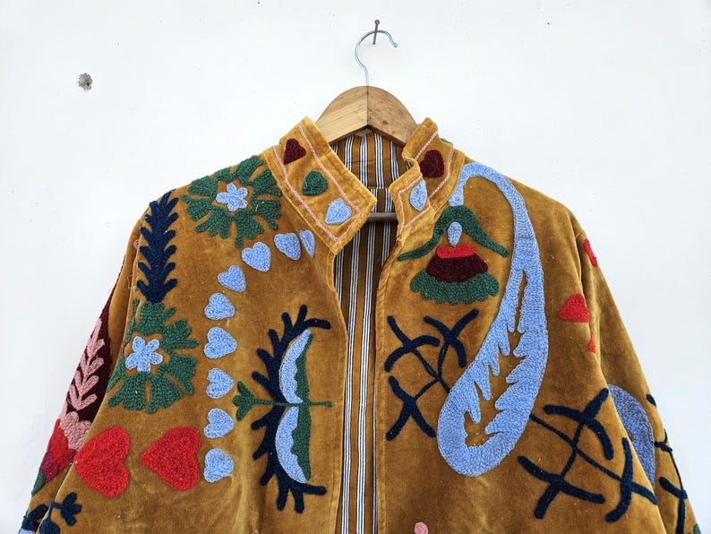 以下が含まれることがあります： マスタード色のジャケットで、複雑な刺繍が施されています。 ジャケットはハイカラーで、花、ハート、抽象的な形など、カラフルな刺繍デザインで飾られています。 裏地は縦縞です。