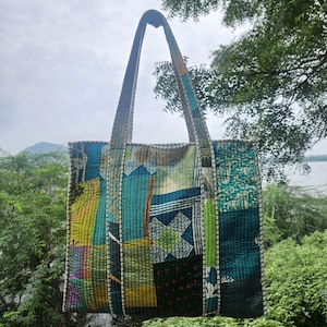 Puede incluir: Un bolso tote acolchado grande con un diseño de patchwork. El bolso presenta una variedad de colores que incluyen verde azulado, verde, amarillo y azul marino. El bolso tiene asas largas y está suspendido frente a un fondo natural.