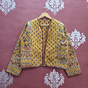 Pode incluir: Casaco acolchoado amarelo com um padrão floral em vermelho, azul e verde. O casaco tem decote em V e é adornado com um tecido vermelho escuro. O casaco está pendurado num cabide de madeira.
