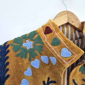 以下が含まれることがあります： マスタード色のジャケット。青、緑、赤の花とハートの刺繍が施されています。ジャケットにはストライプの裏地があり、木製のハンガーに掛けられています。襟はピンクのステッチでトリミングされています。