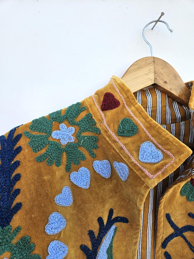以下が含まれることがあります： マスタード色のジャケット。青、緑、赤の花とハートの刺繍が施されています。ジャケットにはストライプの裏地があり、木製のハンガーに掛けられています。襟はピンクのステッチでトリミングされています。