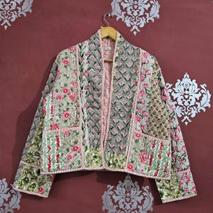 Puede incluir: Chaqueta acolchada con diseño patchwork. Presenta estampados florales en rosa, verde y beige, con forro a rayas rosa y blanco. Cuelga de una percha de madera sobre una pared burdeos.