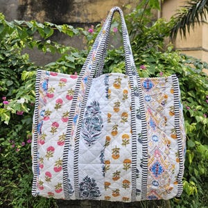 May include: A quilted tote bag with a white background and floral patterns in pink, yellow, and blue. The bag has black and white striped handles and trim. The bag is made of a soft, quilted material.